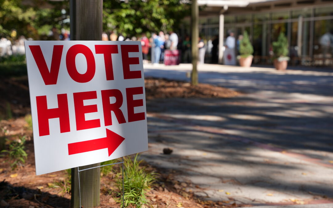 A 'Vote Here' sign with a red arrow points toward a polling place entrance where voters are lined up outside.