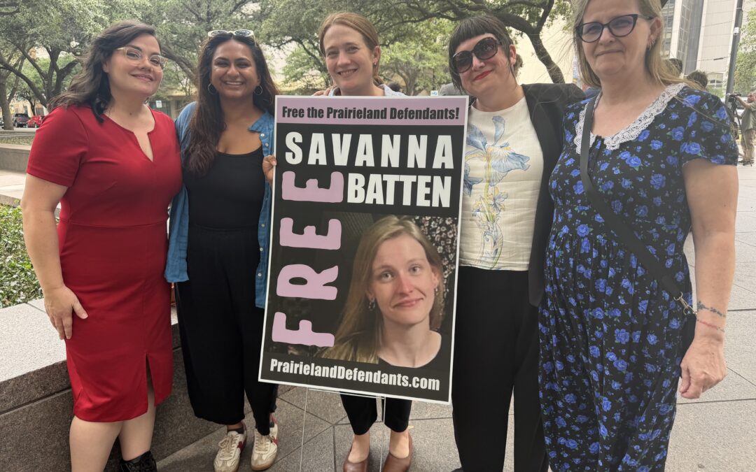 Five people stand together outside a courthouse holding a sign that reads 'Free Savanna Batten — Free the Prairieland Defendants' with a photo of Savanna Batten and the website PrairielandDefendants.com.