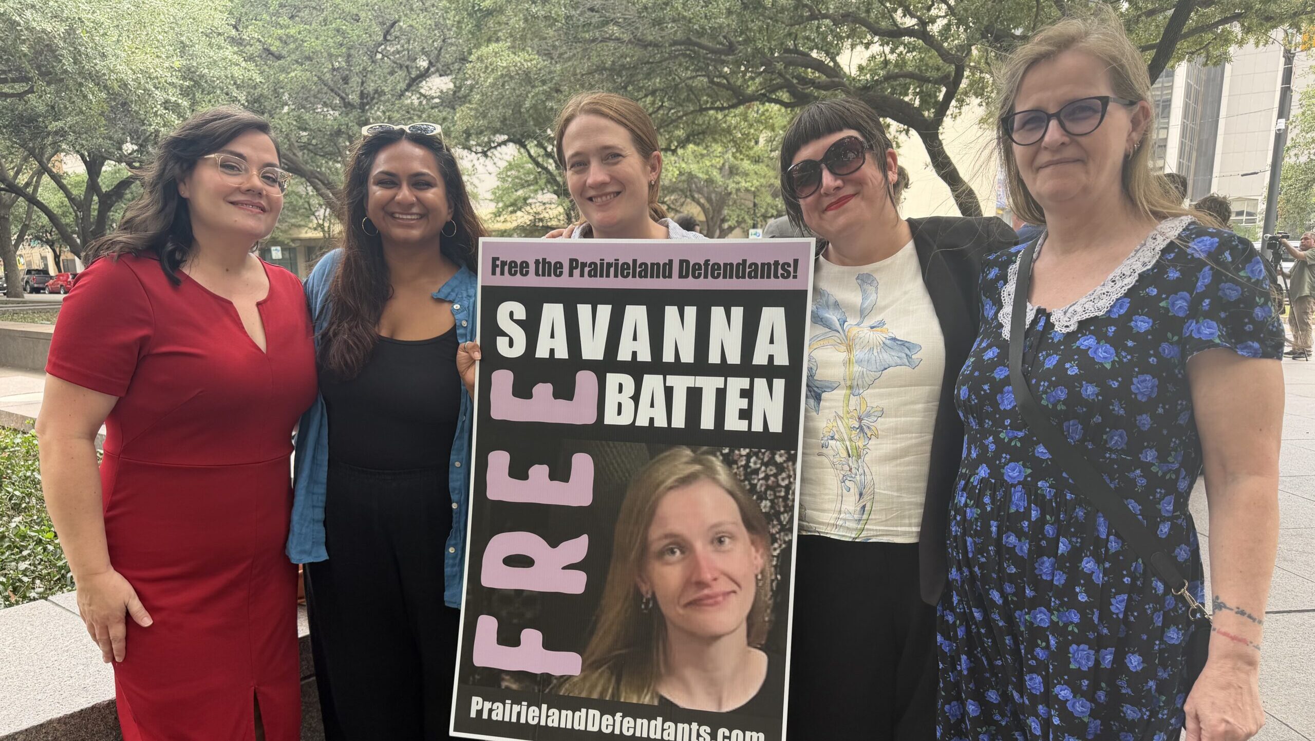 Five people stand together outside a courthouse holding a sign that reads 'Free Savanna Batten — Free the Prairieland Defendants' with a photo of Savanna Batten and the website PrairielandDefendants.com.