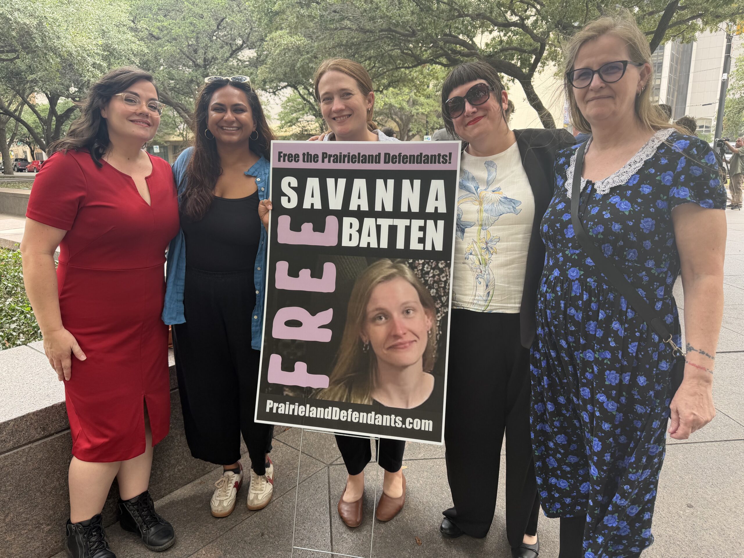 Five people stand together outside a courthouse holding a sign that reads 'Free Savanna Batten — Free the Prairieland Defendants' with a photo of Savanna Batten and the website PrairielandDefendants.com.